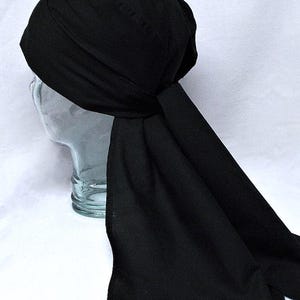 May include: A black head wrap, likely made of a soft fabric, is tied at the back of the head. The wrap covers the head and extends down the back. The head wrap is displayed on a clear glass head form against a white background.