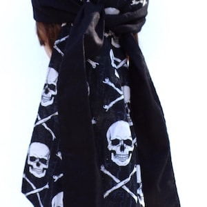 May include: Black head wrap featuring a white skull and crossbones pattern. The head wrap is tied in a knot at the top and has long, solid black fabric hanging down. The fabric is a dark color, and the skulls are detailed.