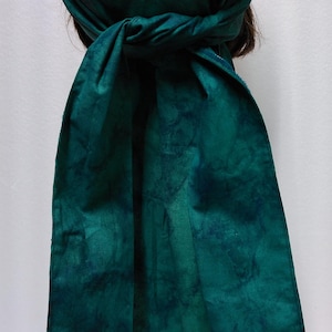 May include: A tie-back head wrap in a teal and dark teal tie-dye pattern. The fabric is wrapped around the head and tied at the back, with long, flowing ties. The head covering is made of soft fabric.