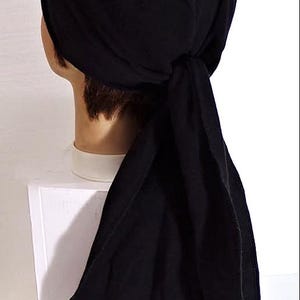 May include: A black head wrap, tied at the back, is displayed on a mannequin head. The fabric appears to be a soft, stretchy material. The head covering is designed to be worn on the head.
