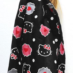 May include: Black headscarf with a repeating pattern of white Hello Kitty faces, pink hearts, and gray cloud shapes. The scarf is tied around a head, with the ends hanging down. White polka dots are scattered across the fabric.
