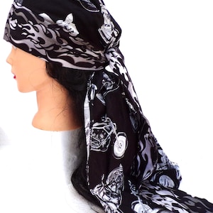 May include: Black head wrap featuring a motorcycle and flame design in white and gray. The head wrap is tied at the back and is displayed on a mannequin head. The fabric has a repeating pattern of motorcycles and flames.