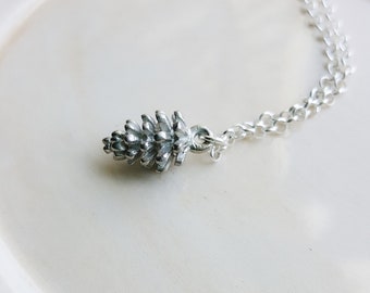 May include: A silver necklace with a pine cone pendant. The chain is a delicate silver chain.