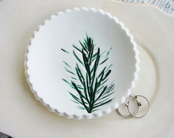 May include: A white ceramic ring dish with a green painted pine tree design. The dish has a scalloped edge.