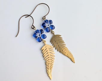 May include: A pair of dangle earrings featuring blue flower beads and gold-colored fern leaf charms. The earrings have a hook closure and are set against a white background. The jewelry is designed for a nature-inspired aesthetic.
