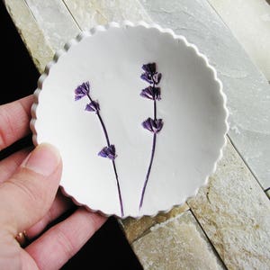 May include: A white ceramic dish with a scalloped edge, featuring two pressed purple lavender flowers.