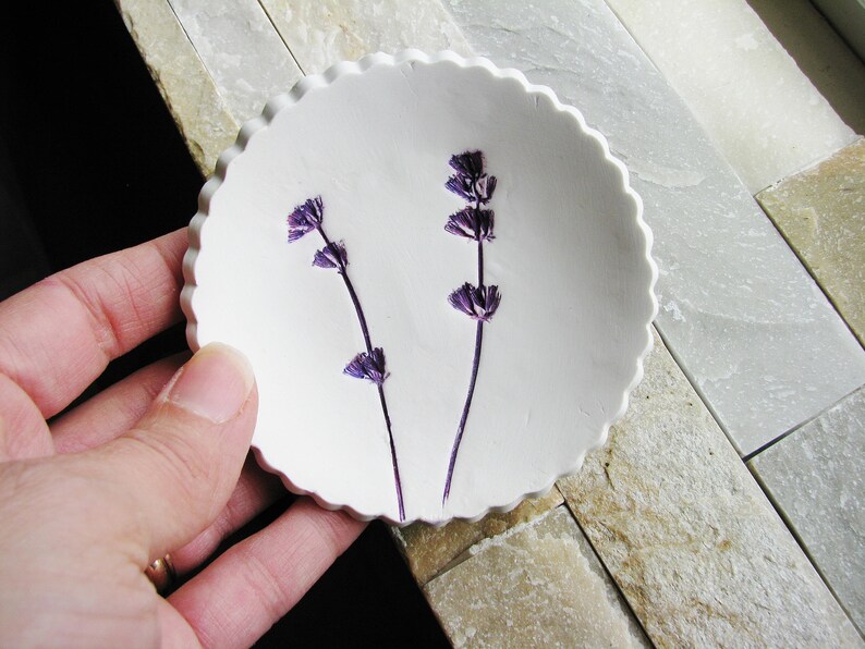 May include: A white ceramic dish with a scalloped edge, featuring two pressed purple lavender flowers.