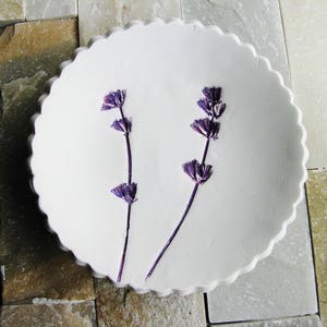 May include: A white ceramic dish with a scalloped edge, featuring two sprigs of dried lavender flowers.