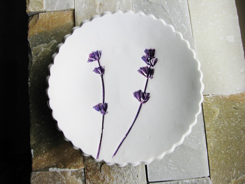 May include: A white ceramic dish with a scalloped edge, featuring two sprigs of dried lavender flowers.