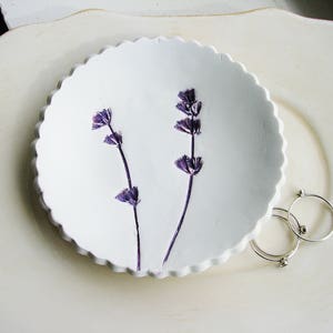 May include: A white ceramic dish with a scalloped edge, featuring two sprigs of dried lavender flowers. The dish is on a white surface.