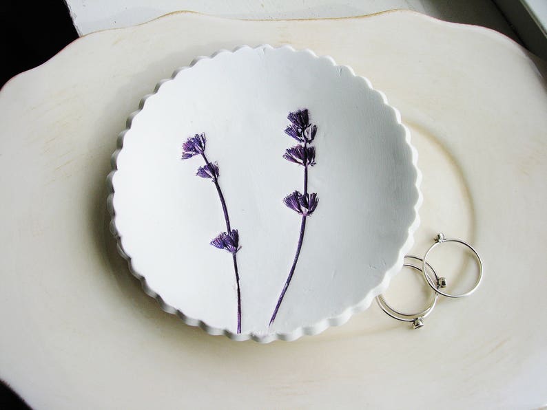 May include: A white ceramic dish with a scalloped edge, featuring two sprigs of dried lavender flowers. The dish is on a white surface.