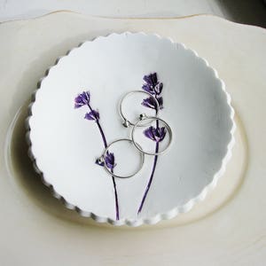 May include: White ceramic dish with a scalloped edge, featuring three silver rings and purple pressed flowers.