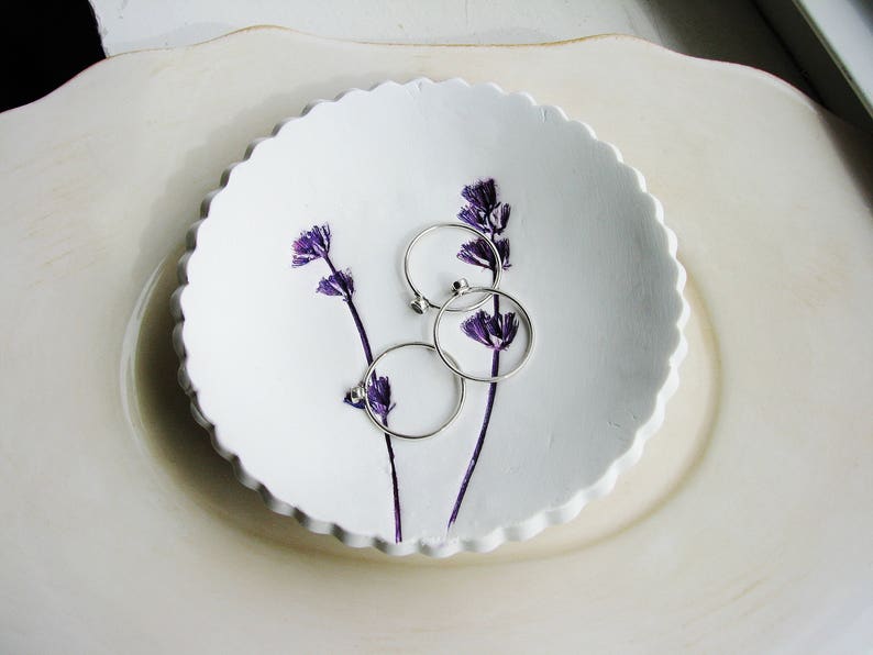 May include: White ceramic dish with a scalloped edge, featuring three silver rings and purple pressed flowers.