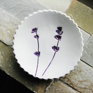 May include: A white ceramic dish with a scalloped edge, featuring two sprigs of purple lavender pressed into the surface.