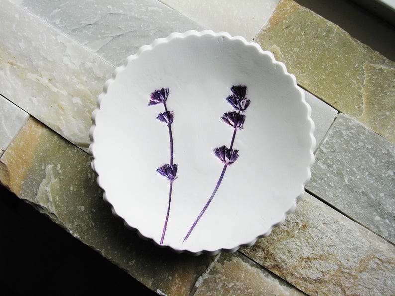 May include: A white ceramic dish with a scalloped edge, featuring two sprigs of purple lavender pressed into the surface.