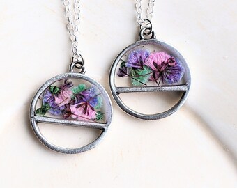 May include: Two silver-toned necklaces with round pendants. Each pendant features a clear resin dome encasing dried flowers in shades of purple, pink, and green. The necklaces are displayed on a white surface, showcasing the delicate floral jewelry.