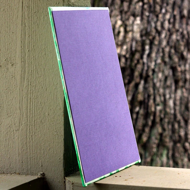 May include: A rectangular, textured, purple object with a white and green border. The object is leaning against a weathered, gray surface, with a blurred tree trunk in the background. The object's surface has a subtle, woven pattern.