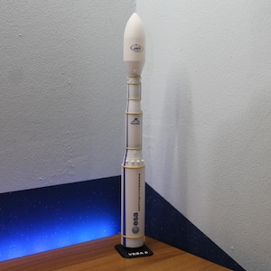 Puede incluir: Un modelo de cohete blanco con detalles azules y dorados. El cohete tiene un cono de nariz blanco con el texto "VEGA" en él. El cohete está sobre una base de madera.