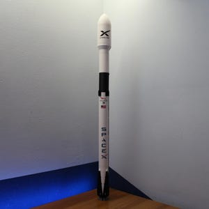 May include: A vertical model of a SpaceX Falcon 9 rocket. The rocket is white with black accents and features the SpaceX logo, the Falcon 9 name, a small American flag, and the Starlink logo on the upper fairing section. It stands in a tall, cylindrical shape.