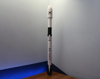 May include: A vertical model of a SpaceX Falcon 9 rocket. The rocket is white with black accents and features the SpaceX logo, the Falcon 9 name, a small American flag, and the Starlink logo on the upper fairing section. It stands in a tall, cylindrical shape.