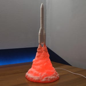 Può includere: Lampada a forma di razzo bianco, con un bagliore arancione brillante alla base, che simula le fiamme. La lampada è su una superficie di legno, con un cavo bianco che ne esce. Lo sfondo è un muro grigio chiaro.