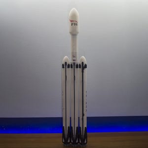 Pode incluir: Um modelo de foguete branco com detalhes em preto e as letras "FH" em vermelho. O foguete tem vários propulsores e um design detalhado, em pé contra um fundo neutro. O modelo é uma representação de um foguete Falcon Heavy.