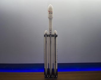 May include: A white model rocket with black accents and the letters "FH" in red. The rocket has multiple boosters and a detailed design, standing upright against a neutral background. The model is a representation of a Falcon Heavy rocket.