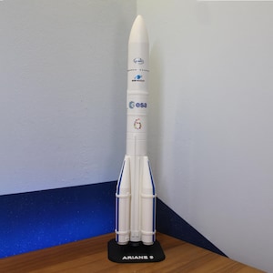 Könnte beinhalten: Ein weißes Modell einer Rakete mit blauen Akzenten und dem Text "Ariane 6" auf einem schwarzen Sockel. Die Rakete trägt das "esa"-Logo und weitere Schriftzüge. Sie steht auf einer Holzoberfläche.