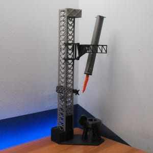 Può includere: Un modello grigio e nero di una torre di lancio di razzi con un razzo e una piattaforma di lancio. Il razzo è grigio con una fiamma rossa. La torre ha un design a traliccio.