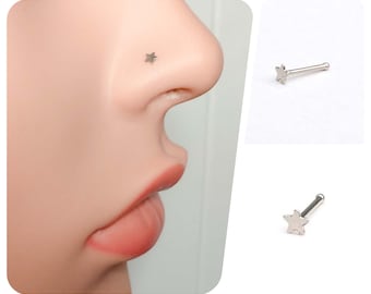 May include: A silver-colored nose stud featuring a small, five-pointed star. The stud is shown in a close-up view, with a side profile of a nose. A second image shows the stud on a white background.