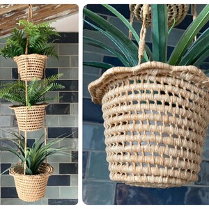 May include: A three-tiered hanging plant holder with woven baskets, each containing a different green plant. The baskets are suspended by natural rope. The background is a blue and grey tiled wall, adding a contrasting backdrop.