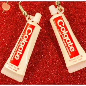 Peut inclure: Boucles d'oreilles fantaisie en forme de tubes de dentifrice Colgate miniatures. Chaque tube blanc porte l'inscription rouge "Colgate" et "FLUORIDE TOOTHPASTE". Les boucles d'oreilles sont suspendues à des crochets argentés.