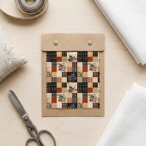 May include: A fabric swatch sample card featuring a patchwork design with alternating squares of cream, black, and burnt orange. The pattern includes plaid, gingham, and botanical floral illustrations in a grid layout, secured by two metal rivets.