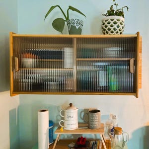 May include: A light wood cabinet with ribbed glass doors is mounted on a light blue wall. The cabinet has two shelves and is filled with various items. Two potted plants sit on top of the cabinet. Below, a small table holds mugs and a teapot.