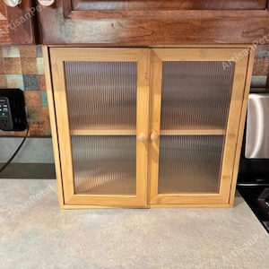 May include: A light-colored wooden cabinet with two doors and clear, textured glass panels. The cabinet has a simple design with wooden knobs and is likely for kitchen storage. The cabinet is sitting on a countertop.