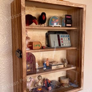 May include: A wooden display cabinet with a glass front, showcasing various vintage collectibles. The cabinet features multiple shelves holding items like figurines, a snow globe, and a vintage radio. The cabinet is made of light brown wood.