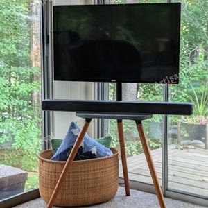 May include: A black flat-screen TV mounted on a modern, dark gray stand with wooden legs. A soundbar is positioned below the TV. A woven basket with pillows sits on the floor. The stand is on a light-colored carpet.