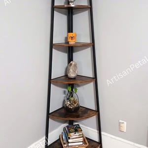 May include: A corner bookshelf with five triangular shelves. The shelves are a dark wood color, supported by a black metal frame. The shelves are decorated with a variety of items, including a small owl figurine, a mug, a rock, a glass terrarium, and books.