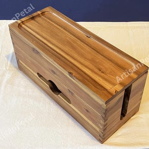 May include: A rectangular wooden cable box with a natural wood finish. The box has a lid with a recessed area and slots on the sides for cable management. The box is made of light brown wood and sits on a white surface.