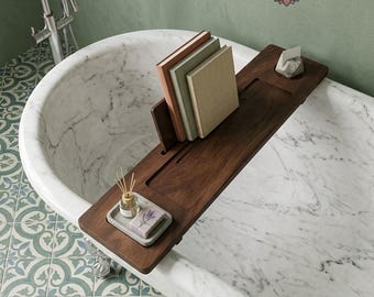 Puede incluir: Una bandeja de madera marrón oscuro para bañera colocada sobre una bañera de mármol blanco. La bandeja cuenta con un soporte ajustable para libros con tres libros, una ranura rectangular y una pequeña bandeja gris con difusor y jabón.