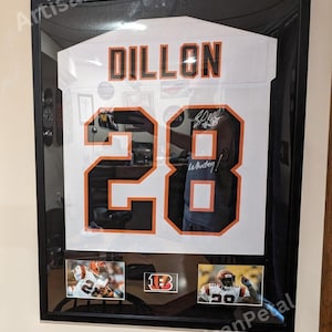 May include: Framed football jersey with the name "DILLON" and the number "28" in orange on a white background. The jersey is signed and includes two action photos of a football player and a team logo. The frame is black.