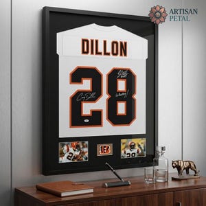 May include: A framed white sports jersey with the name DILLON and the number 28 in black with orange outlines. The jersey features two black ink signatures on the number 28. Below the jersey are two small photographs of a player and a team logo.