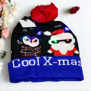 May include: Black knitted hat with a blue brim and a red pom-pom. The hat features a light-up owl and Santa Claus design, with the words "Cool X-mas" in white on the brim. A blank tag hangs from the hat.