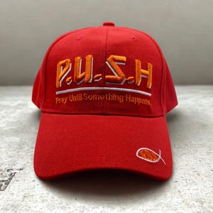 以下が含まれることがあります： オレンジと白で「P.U.S.H. Pray Until Something Happens」と刺繍された赤い野球帽。つばには小さな魚のシンボルが刺繍されています。