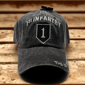 May include: A dark gray baseball cap with the text "1ST INFANTRY" embroidered in white above a white-bordered black shield with the number "1". The cap has a curved brim and the text "THE BIG" on the side.