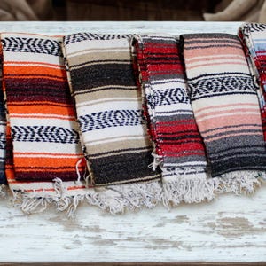 May include: A collection of eight folded woven blankets featuring various color combinations including red, blue, orange, tan, black, and white. Each blanket displays a repeating geometric pattern and is finished with a fringed edge at the bottom.