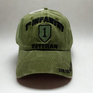 May include: Olive green baseball cap with embroidered text and a shield emblem. The cap reads "1st INFANTRY VETERAN" in black thread. The brim has the text "THE BIG RED ONE".