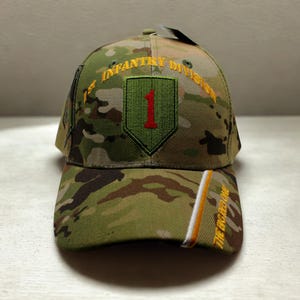 May include: Camouflage baseball cap featuring the text "1st Infantry Division" in yellow embroidery. The cap has a green patch with a red "1" and the words "THE BIG RED ONE" in yellow.