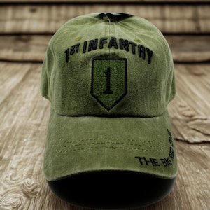 May include: An olive green baseball cap with the text "1st Infantry" arched above a shield with the number "1". The phrase "THE BIG RED ONE" is printed on the side of the cap. The cap is displayed on a black form.