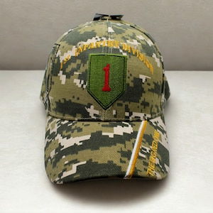 May include: A camouflage baseball cap with the text "1st Infantry Division" and a patch featuring a green shield with a red "1". The cap has a curved brim and a white and yellow stripe.